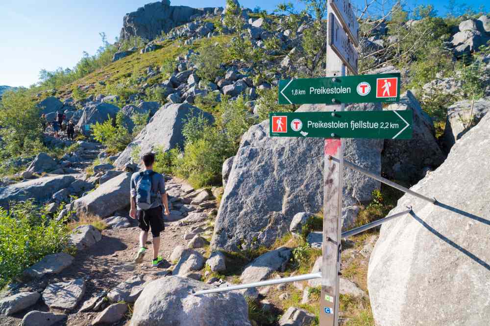 Hiker on rocky path passing green signs pointing to Preikestolen and the mountain lodge.