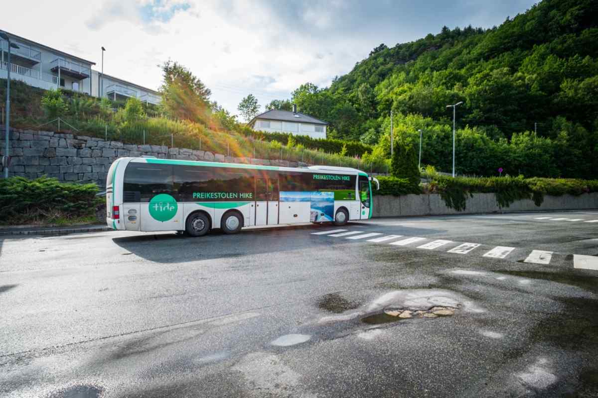Getting around Preikestolen by bus Bus departing toward the Preikestolen hike in Norway, parked along a quiet road with green hillsides and residential buildings nearby.