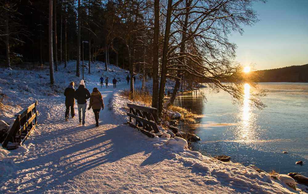 People walking along a snowy riverside path at sunset in Oslo’s winter forest.