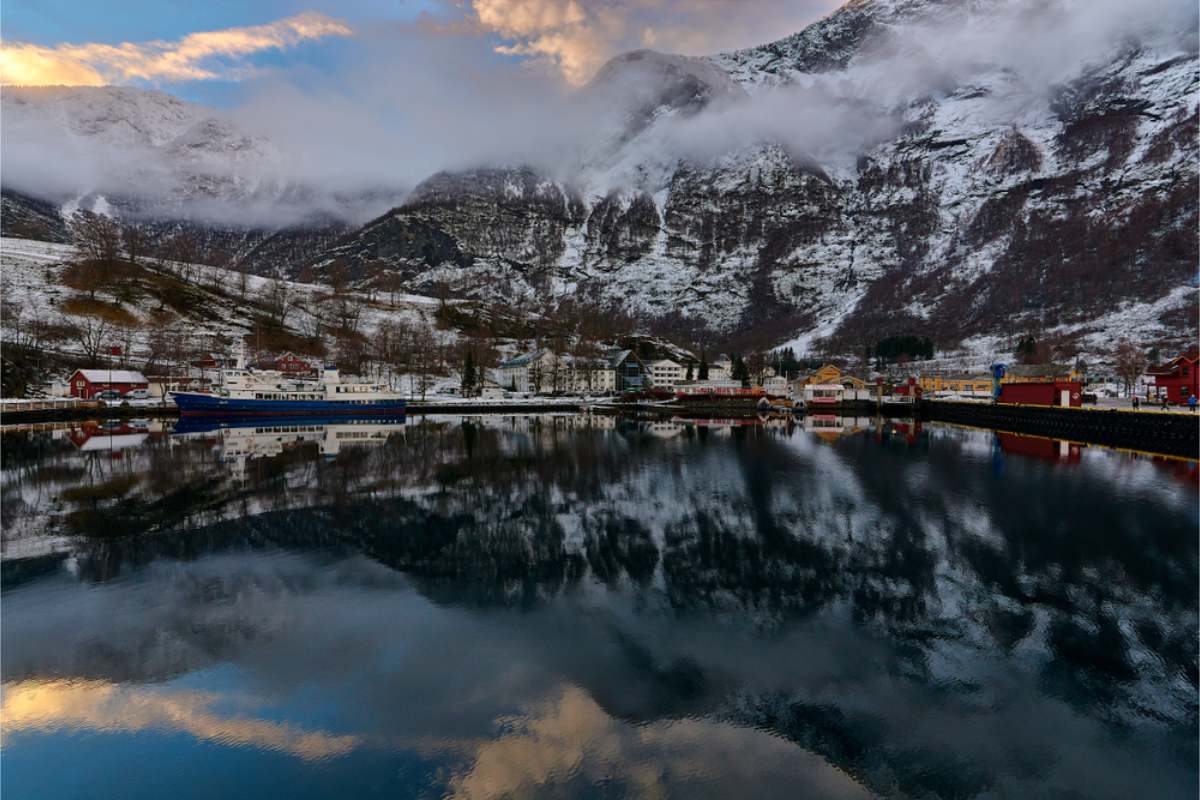A calm fjord water beneath snow-dusted peaks in Norway Small waterside village with red and white houses and a moored ferry, reflected in calm fjord water beneath snow-dusted peaks