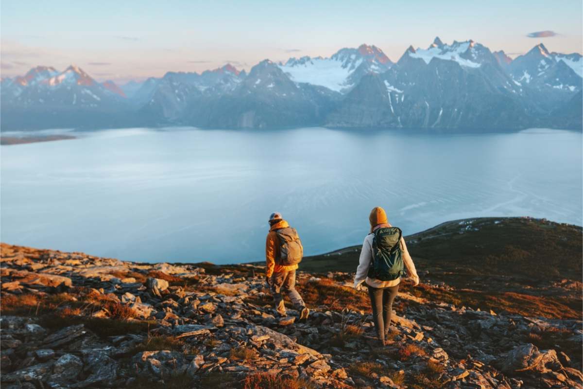 A wide fjord and sharp snow-covered peaks in Norway Two hikers with backpacks walking down a rocky slope at sunrise, overlooking a wide fjord and sharp snow-covered peaks