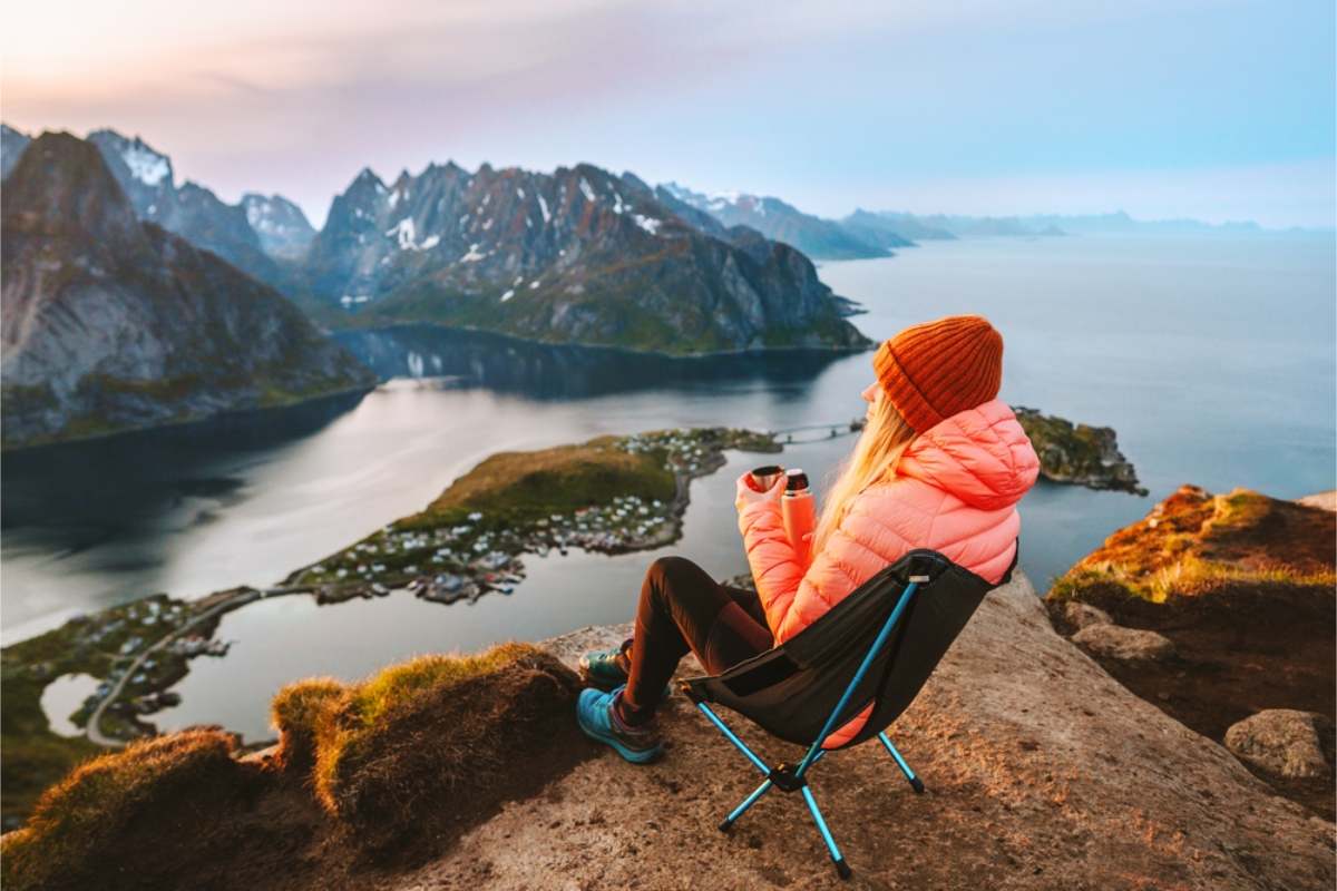 A fishing village in Norway Woman in a pink jacket sitting in a folding chair on a clifftop with a thermos, looking down at a fishing village between fjords