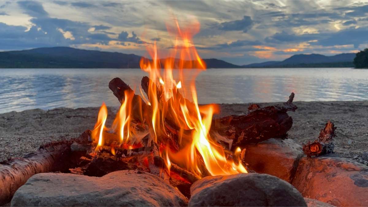 A lake and dark hills in Norway Open campfire burning between stones on a pebble beach, with a lake and dark hills behind under an evening sky