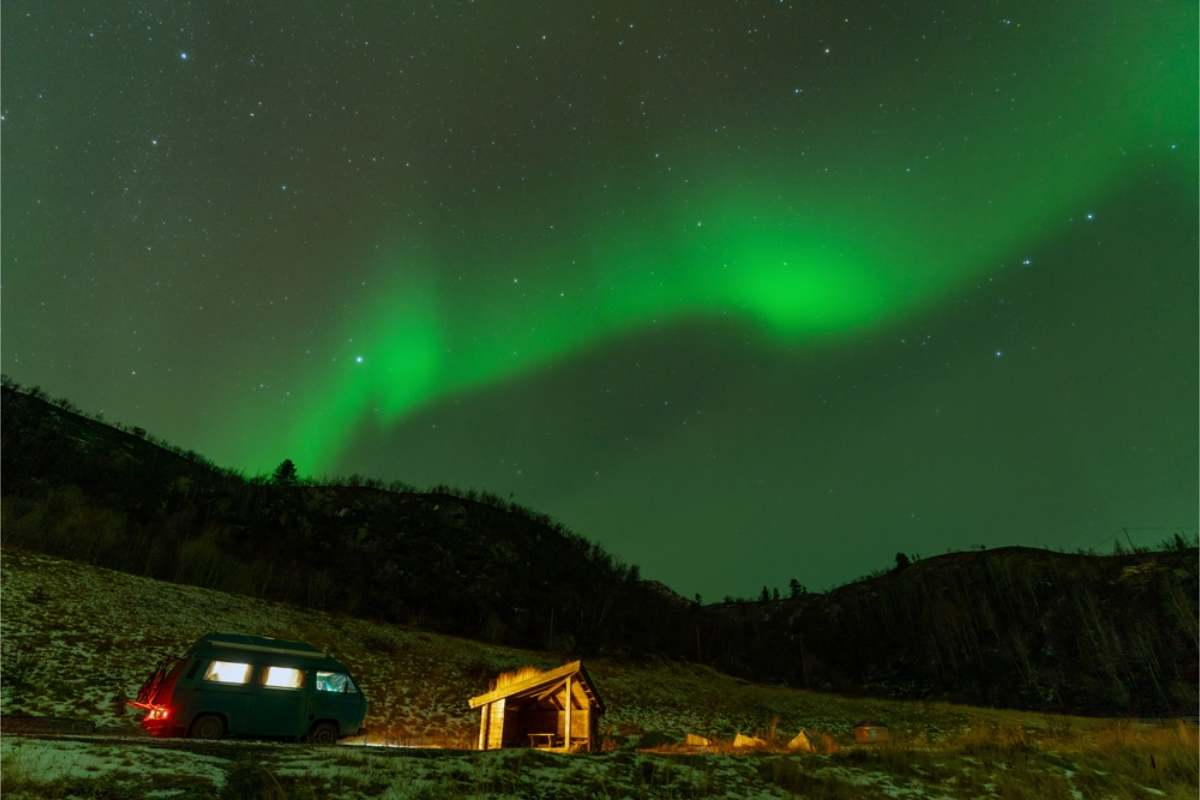 Northern lights in Norway Green aurora across a starry sky over a van parked beside a small wooden shelter on a snow-patched hillside