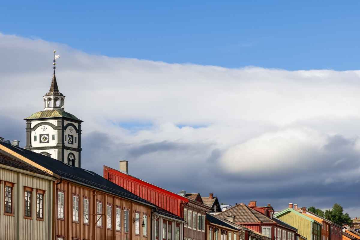 Hyttklokka in Roros The Hyttklokka clock tower in Røros above a row of traditional wooden buildings under a blue sky with dramatic clouds.