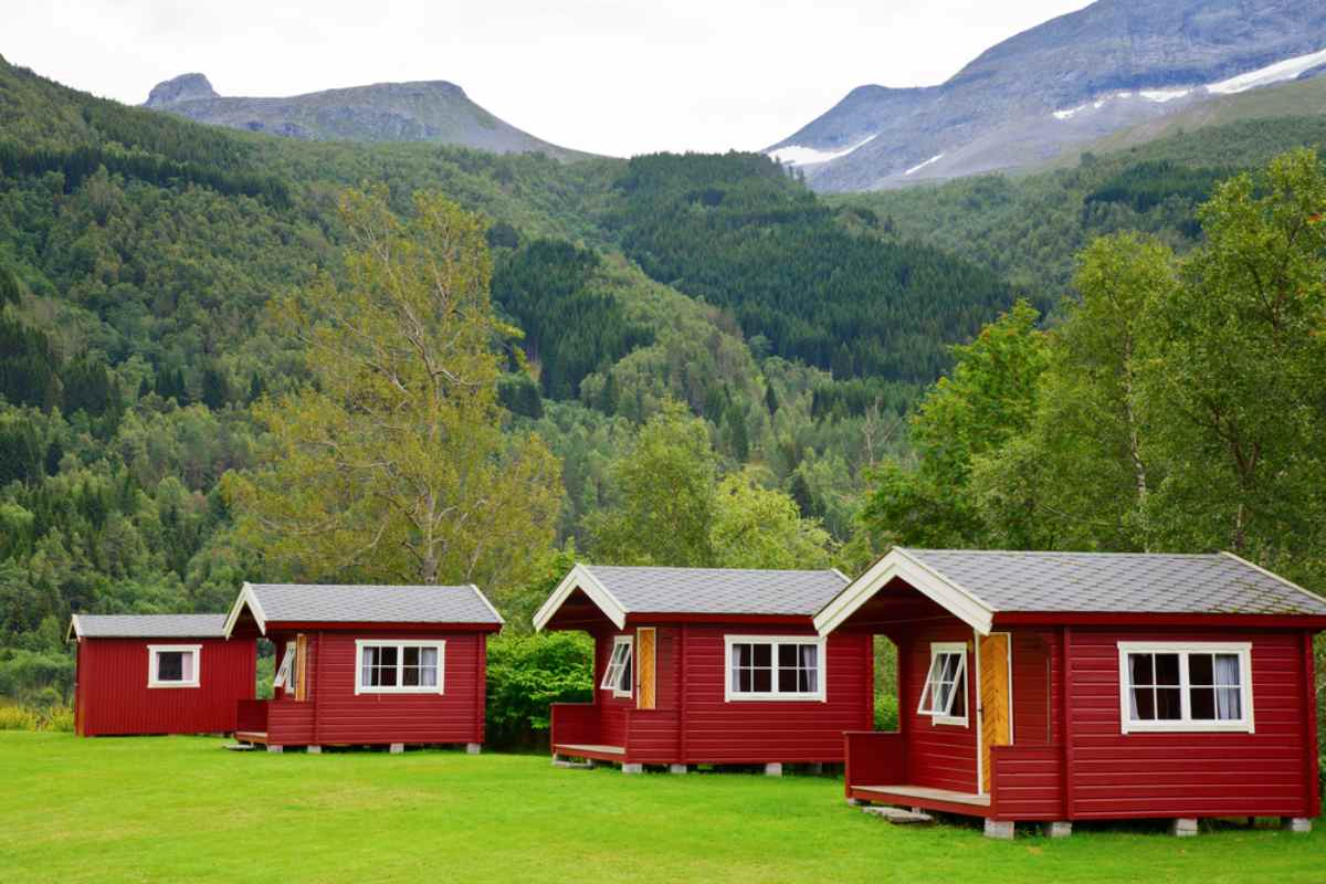 Red wooden camping cabins with white trim on a grassy field, forested hills and mountains in the background.