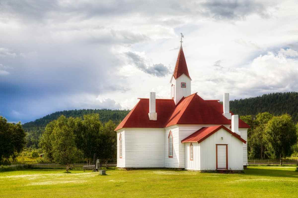 White wooden church with red roof surrounded by green trees and grassy lawn under a partly cloudy sky.