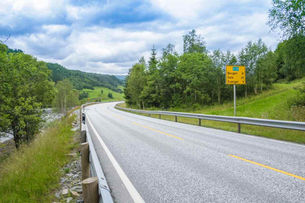 Overview of Norway's E16 road with green vegetation surrounding it and a sign showing the nearest towns on the right side of the road.