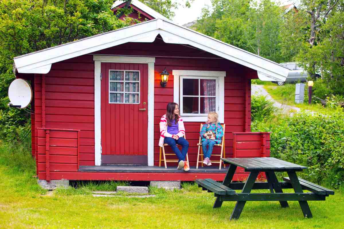 Two girls, an older brunette and a little blonde, enjoy the view from a campsite inside the porch of a reddish wooden cabin, sitting on chairs at a table.
