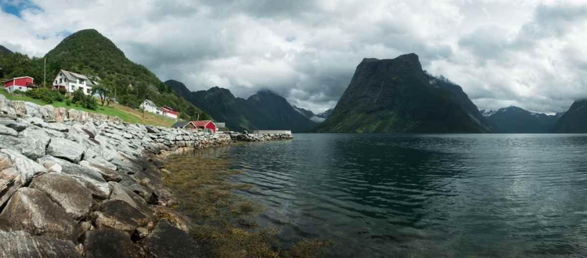 Waterfront village and rocky shoreline on Hjørundfjord, steep mountains under cloudy skies near Ålesund, Norway.