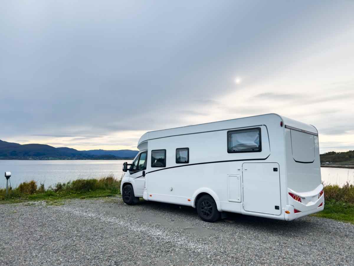White motorhome parked at a waterfront campsite with calm water and distant hills near Ålesund, Norway.