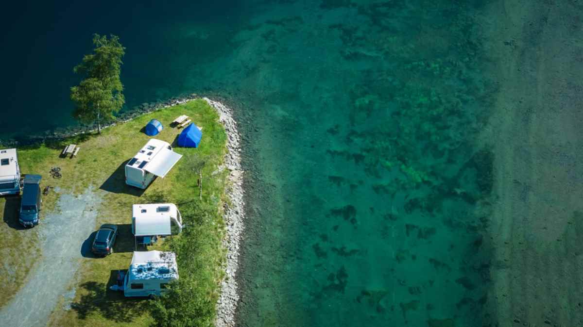 Aerial view of RVs and tents on a narrow lakeside campsite with clear turquoise water.