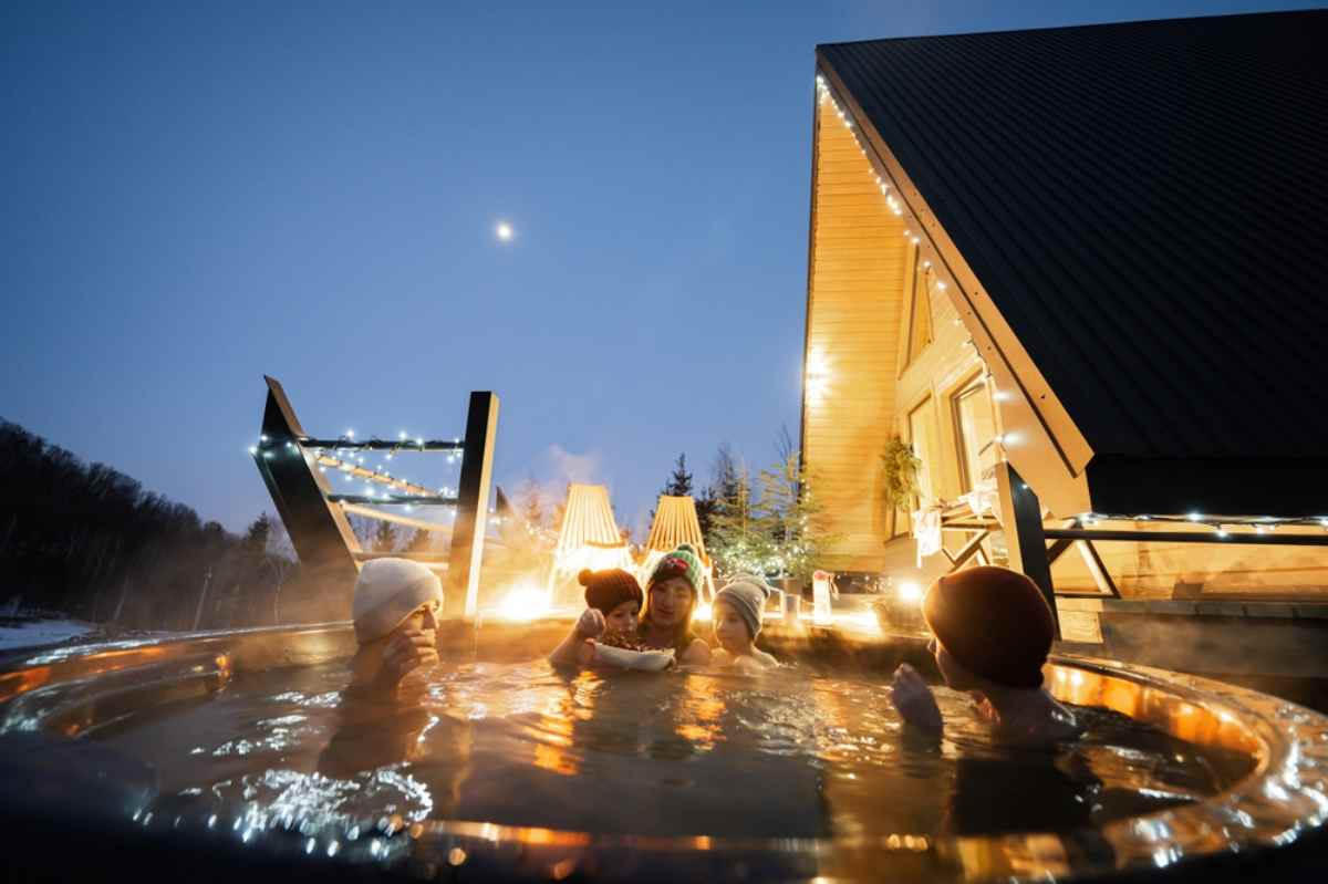 Family relaxing in a steaming outdoor hot tub beside a cabin strung with lights at dusk.