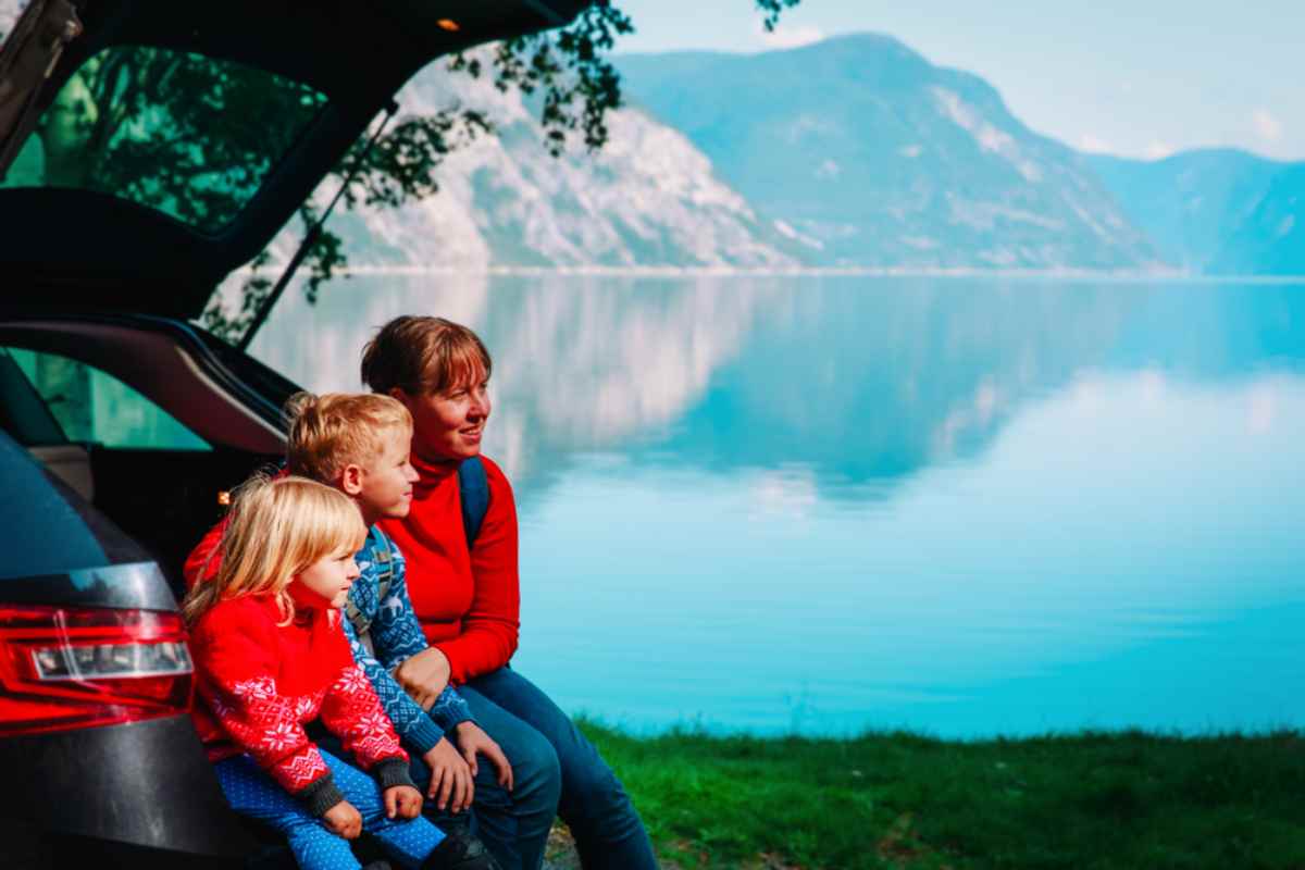 Mother and two kids sitting on a car’s open tailgate by a calm fjord and mountains.