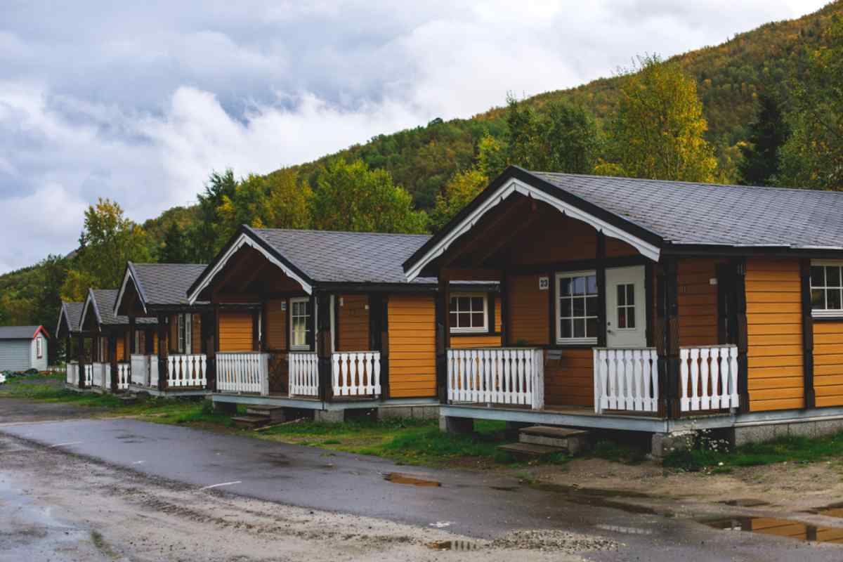 Campsites in Northern Norway Row of cozy wooden camping cabins with white railings in Norway, puddled driveway and forested hillside behind under overcast autumn skies.