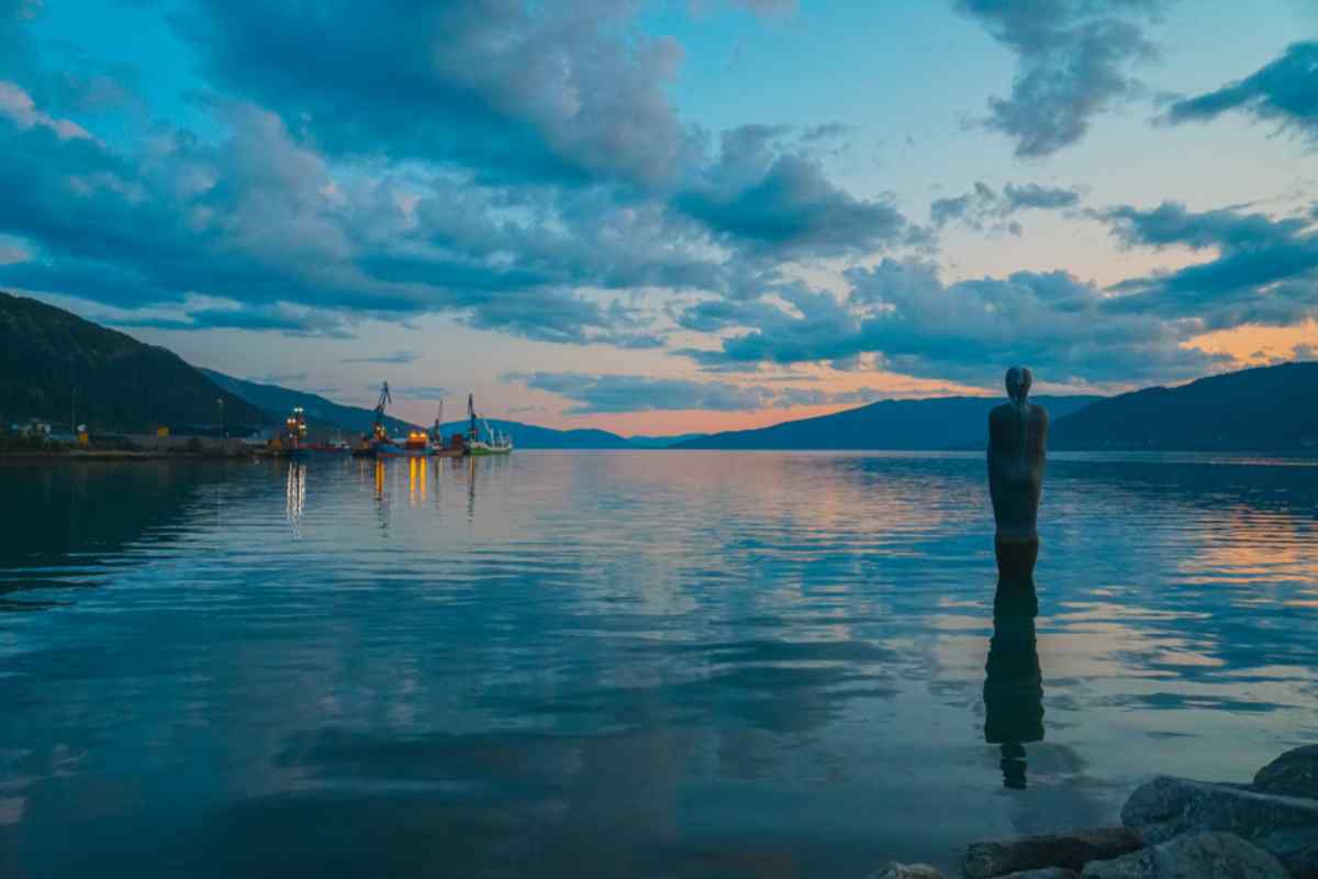 Mo i Rana, Norway Havmannen statue stands in the fjord at Mo i Rana, calm evening water and harbor cranes in the distance beneath blue clouds and sunset light.