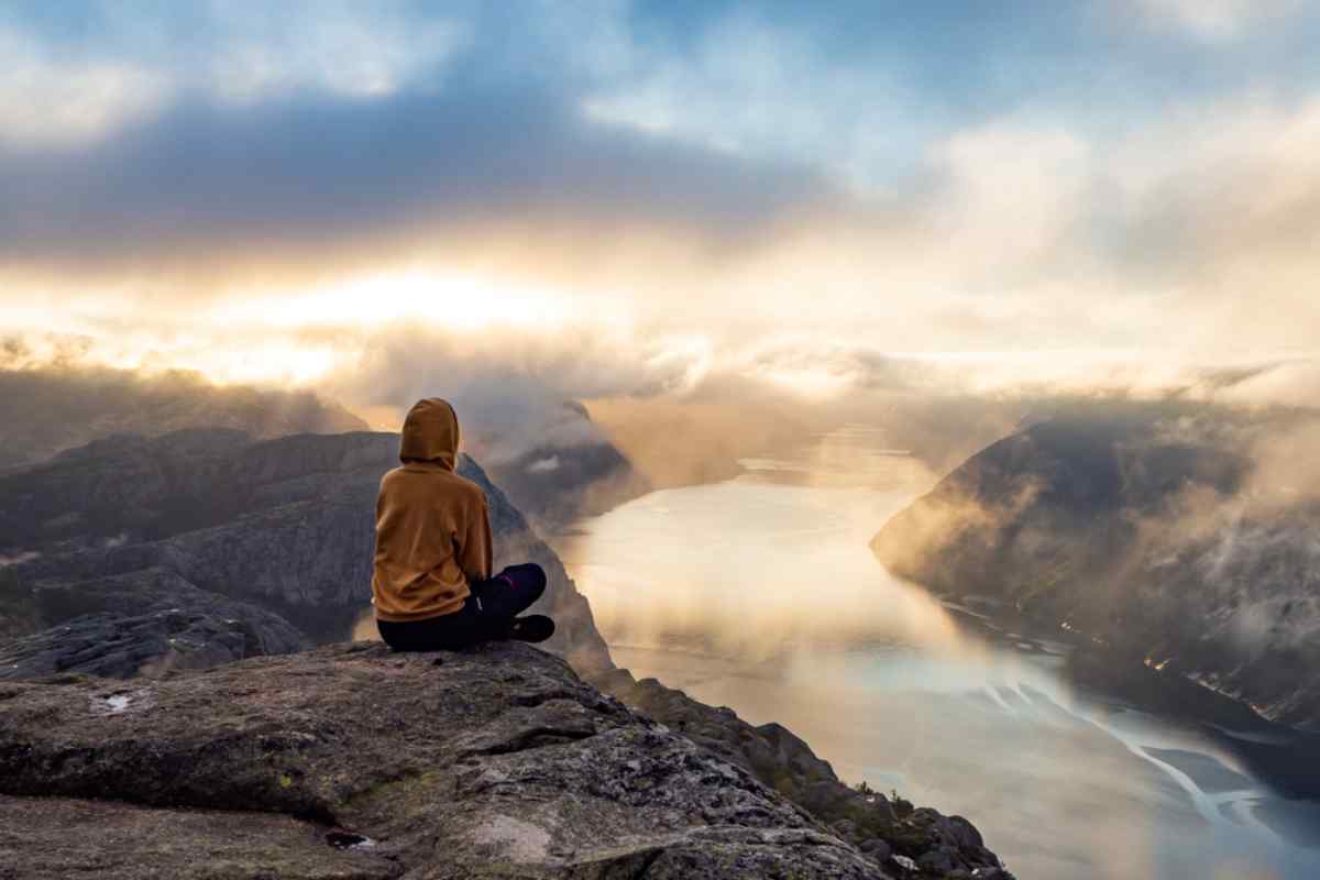 Preikestolen in Norway Hiker in a hooded jacket sits on the edge at Preikestolen, watching sunrise light spill over Lysefjord’s cliffs and winding water below.