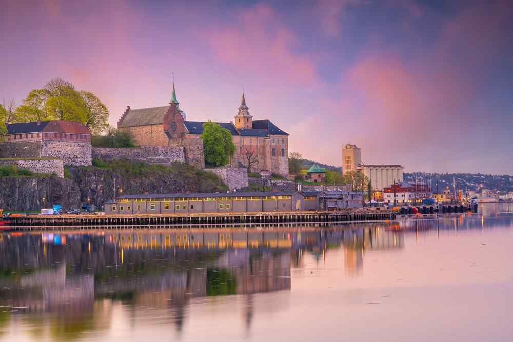 Oslo, Norway Akershus Fortress and Oslo waterfront reflected in calm water at sunset.