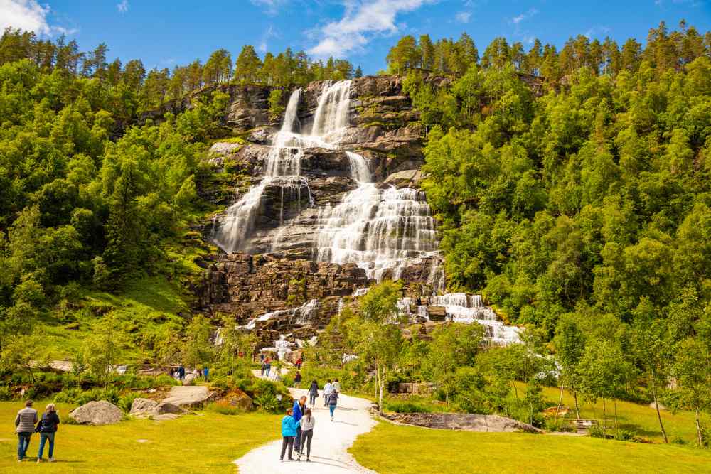 Tvindefossen Waterfall Visitors walking toward Tvindefossen waterfall surrounded by green forest in Norway.