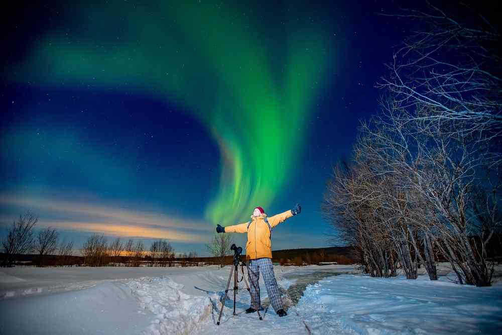Best time to see the northern lights in Lofoten Traveler standing beside a camera tripod under vivid green northern lights over a snowy landscape in northern Norway.