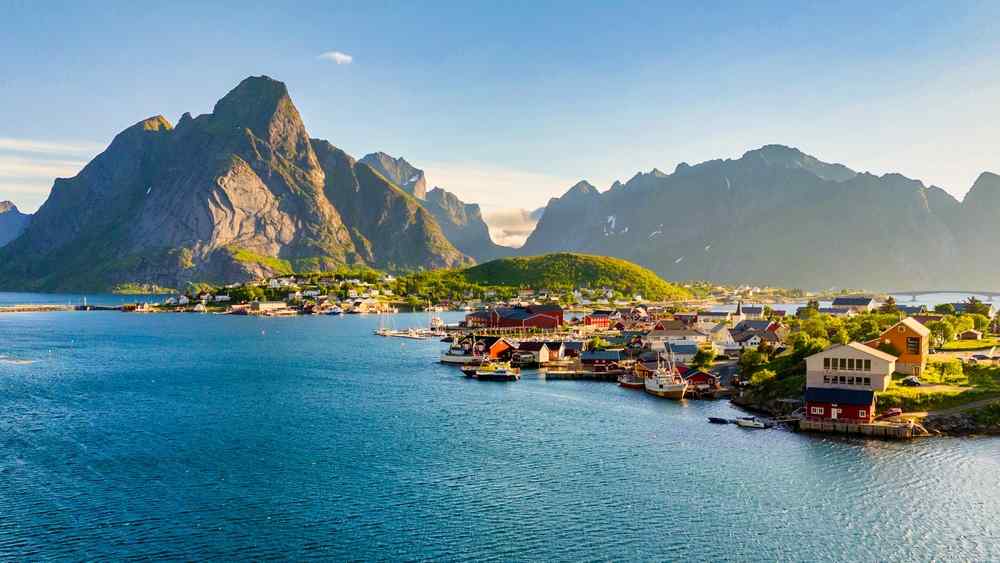 Best time to see the northern lights in Lofoten Panoramic view of a coastal village in the Lofoten Islands surrounded by blue water and dramatic rugged mountains.