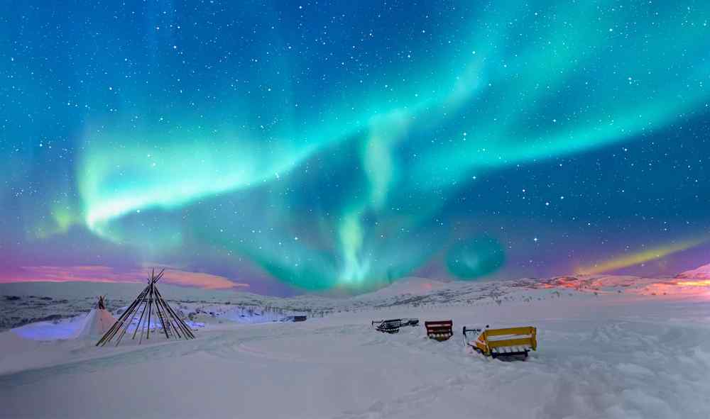 Vibrant green northern lights stretching across a starry sky above a snowy Arctic landscape with traditional wooden structures.