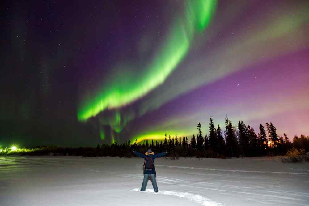 Traveler standing with arms raised beneath vivid green northern lights over a snowy landscape surrounded by dark pine trees.